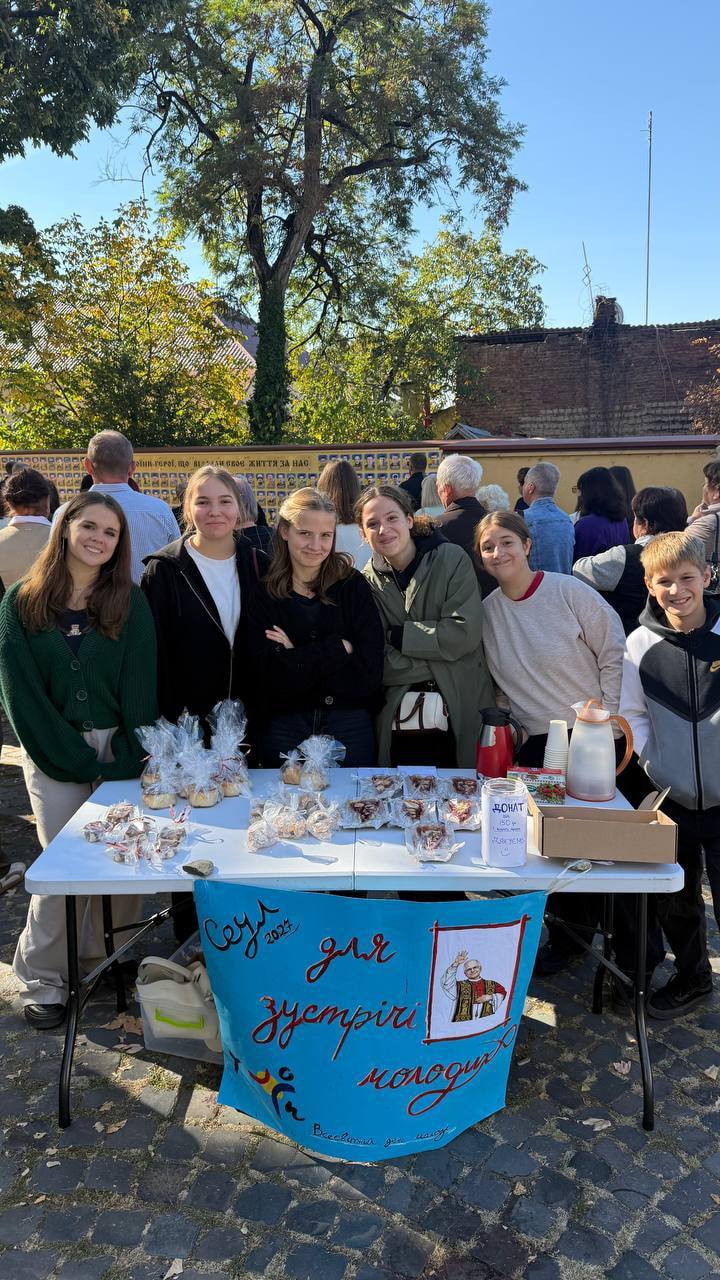 Молодь біля столу благодійного ярмарку з випічкою в пакетиках і банером «Сеул 2027 для зустрічі молоді»
